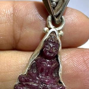 Op de afbeelding: Een hanger met een donkerrood, uit edelsteen gesneden figuur van een zittende godheid in een zilveren zetting. De zilveren omlijsting bevat een sierlijk, driehoekig oogje met krulpatronen aan de bovenzijde dat de gepolijste steen vasthoudt.