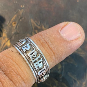 May include: A silver ring with an inscription that reads "ALPHA" in a stylized font. The ring is worn on a finger.