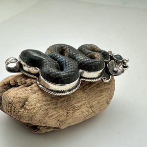 May include: A handcrafted silver and stone snake pendant. The pendant features a detailed snake design with a textured, dark gray body and silver accents. The snake is coiled and rests on a piece of driftwood.