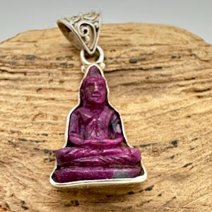 May include: A pendant featuring a carved deep magenta ruby gemstone in the shape of a seated Buddha figure. The gemstone is set within a silver frame and includes a decorative silver bail with intricate scrollwork at the top of the piece.