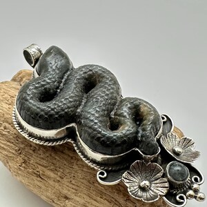 May include: A dark green, textured snake pendant with a silver-colored border and floral accents. The snake is coiled, and the pendant has a loop for a chain. The floral accents include three flowers and a small, round stone.