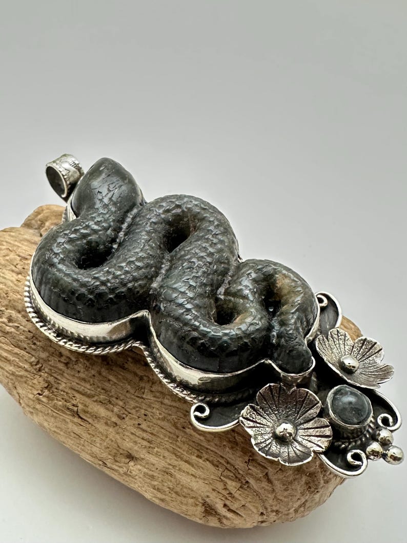 May include: A dark green, textured snake pendant with a silver-colored border and floral accents. The snake is coiled, and the pendant has a loop for a chain. The floral accents include three flowers and a small, round stone.