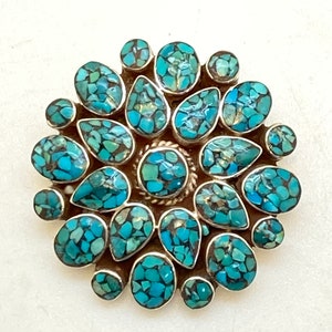 Puede incluir: Un colgante de plata con un diseño de piedra turquesa. La turquesa es un mosaico de pequeñas piezas irregulares en tonos azules y verdes.