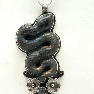 May include: A silver pendant necklace featuring a stylized snake design. The snake is dark gray with iridescent highlights, set within a silver frame. The pendant includes floral and gemstone accents, suspended from a delicate chain.