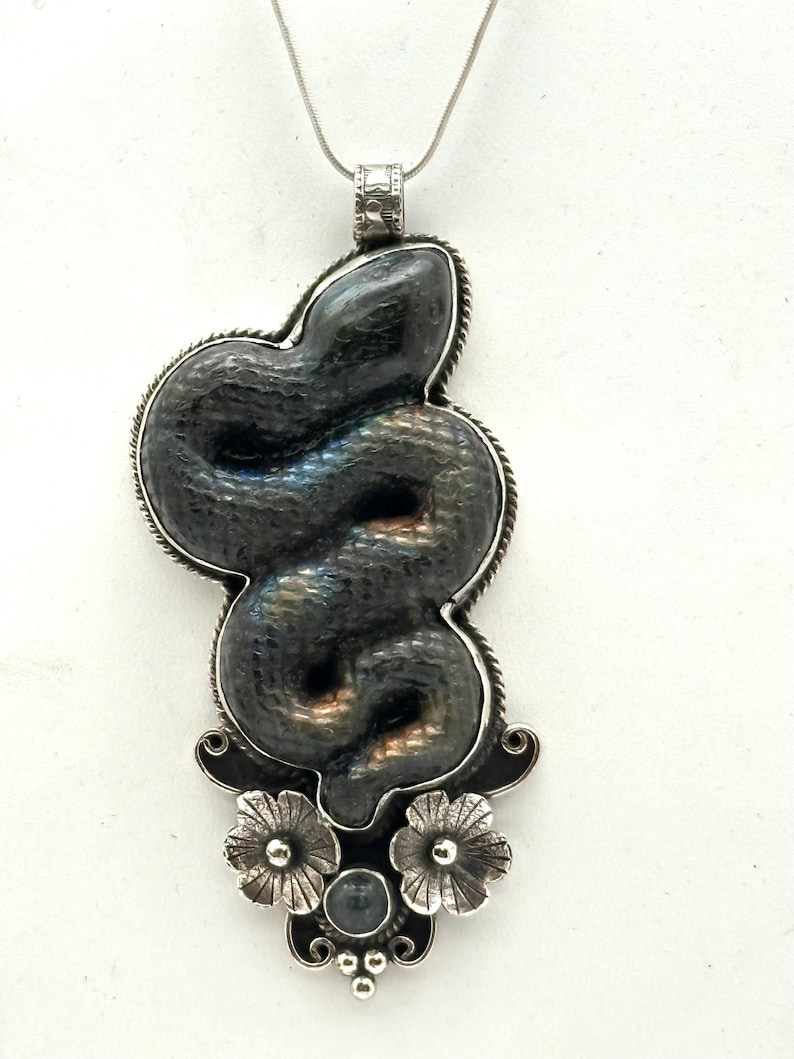 May include: A silver pendant necklace featuring a stylized snake design. The snake is dark gray with iridescent highlights, set within a silver frame. The pendant includes floral and gemstone accents, suspended from a delicate chain.