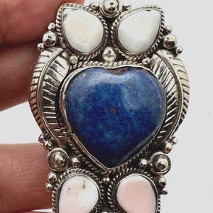 May include: A silver brooch featuring a central heart-shaped blue lapis lazuli stone, flanked by two white teardrop stones at the top and two smaller teardrop stones in white and pale pink at the bottom, all set in ornate, polished silver metalwork.