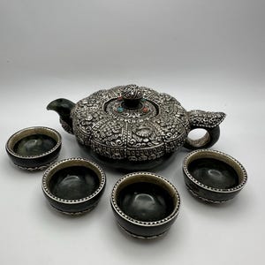 Könnte beinhalten: Eine verzierte silberne Teekanne mit aufwendigen Details und einem passenden Satz von vier kleinen, dunkel gefärbten Schalen. Die Teekanne hat einen dekorativen Deckel mit farbigen Akzenten. Die Schalen und die Teekanne sind auf einer weißen Oberfläche angeordnet.