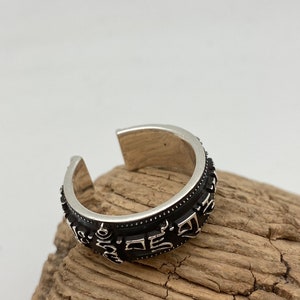Könnte beinhalten: Ein silberfarbener Ring mit einem schwarzen Band, das mit eingravierten Symbolen versehen ist. Der offene Ring liegt auf einem Stück Treibholz. Das Design des Rings ist detailliert und aufwendig.