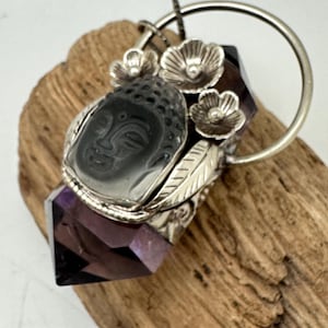 May include: A silver pendant featuring a carved Buddha face set in a clear amethyst crystal point. The pendant is adorned with three silver flower accents and a silver loop for hanging.
