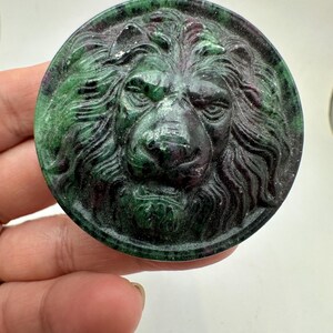 May include: A circular stone carving featuring a detailed lion head in relief. The piece is crafted from a mottled green and dark red mineral, showcasing a smooth, polished surface with natural color variations throughout the lion's mane and face.