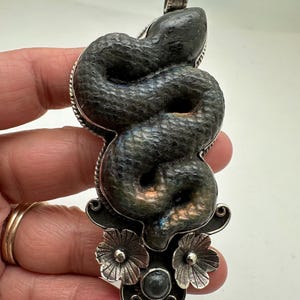 May include: A silver pendant featuring a coiled snake design. The snake is dark gray with iridescent blue and green hues. The pendant has a floral design at the bottom and a decorative bail at the top. The pendant is approximately 3 inches long.
