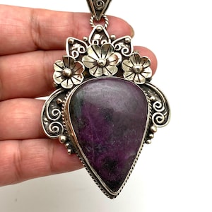 May include: A large, ornate pendant with a teardrop-shaped, deep purple stone. The pendant is framed in silver-toned metal with floral and scrollwork details. The bail is a decorative triangle shape.