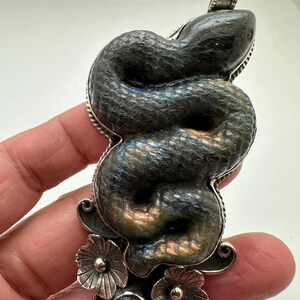 May include: A silver pendant featuring a coiled snake design. The snake is a dark gray color with iridescent blue and gold hues. The pendant has a floral design at the bottom and a loop at the top for a chain.