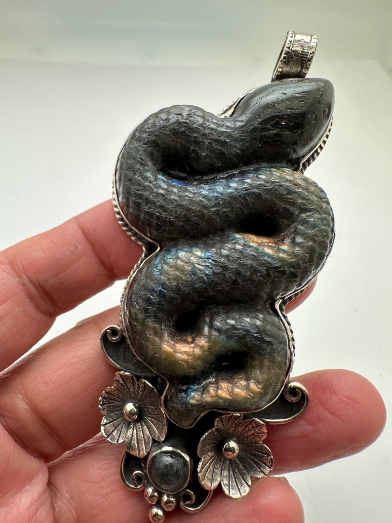 May include: A silver pendant featuring a coiled snake design. The snake is a dark gray color with iridescent blue and gold hues. The pendant has a floral design at the bottom and a loop at the top for a chain.