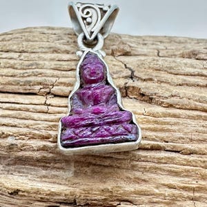 May include: A pendant featuring a deep magenta-colored carved stone figure in a seated position, encased in a silver-toned metal setting. The setting includes a decorative triangular bail with scrollwork patterns at the top of the pendant.