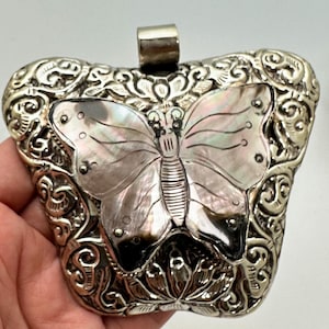 Puede incluir: Colgante plateado con diseño de mariposa. La mariposa tiene alas iridiscentes y está engastada en un marco decorativo. El colgante tiene un lazo en la parte superior para una cadena.