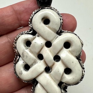 May include: A pendant featuring a white, carved, knot-shaped centerpiece set within a silver frame. The design includes a circular, dark, polished stone at the top bail, surrounded by a twisted silver rope border that outlines the entire knot shape.
