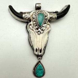 May include: A pendant featuring a carved white bone bull skull with dark horn tips, accented by silver metal filigree and a teardrop-shaped turquoise stone on the forehead. A matching teardrop turquoise stone hangs from the base of the skull.