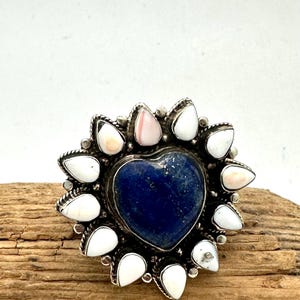 Puede incluir: Un anillo de plata con una piedra central azul oscuro en forma de corazón, rodeada por un halo de piedras blancas y rosa pálido en forma de lágrima. El engaste metálico tiene una textura de cuerda trenzada alrededor del corazón central.