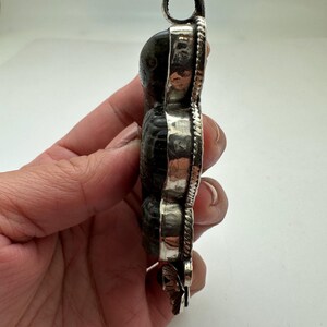 May include: A pendant featuring a dark, textured stone set in a silver frame. The pendant has a loop at the top for a chain and a decorative floral element at the bottom. The stone appears to be a fossil or a similar natural material.