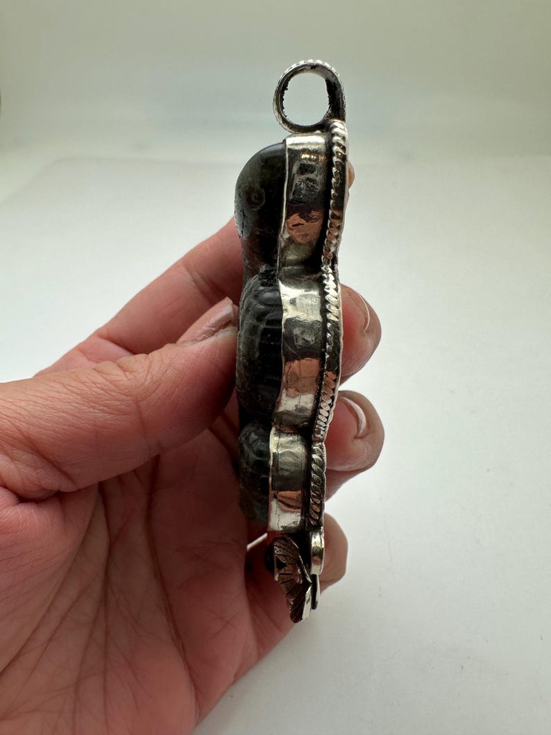 May include: A pendant featuring a dark, textured stone set in a silver frame. The pendant has a loop at the top for a chain and a decorative floral element at the bottom. The stone appears to be a fossil or a similar natural material.