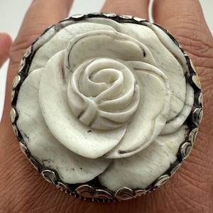 Op de afbeelding: Een grote, ivoorkleurige ring met een gebeeldhouwd rozenontwerp. De roos heeft gedetailleerde bloemblaadjes en is gezet in een zilverkleurige band met decoratieve accenten. De ring is een statement piece.