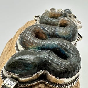 May include: A detailed pendant in the shape of a snake, crafted from a dark, iridescent stone with silver accents. The snake's body is coiled, and the pendant has a textured surface. The silver setting features intricate detailing, including a rope-like border and decorative elements.