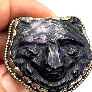May include: A rectangular, carved amethyst pendant featuring a stylized bear face. The bear's features are detailed with a dark purple hue, set within a gold-toned, beaded frame. The pendant is held by a hand.