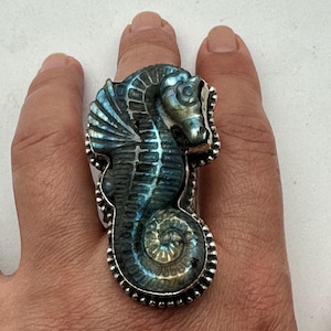 May include: A large statement ring featuring a carved labradorite gemstone in the shape of a seahorse. The stone displays iridescent blue and gold flashes set within a polished silver frame with a beaded edge detail along the entire perimeter.