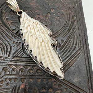 May include: A silver pendant in the shape of a wing with a detailed design. The wing is white and has a textured surface. The pendant is hanging from a silver chain.