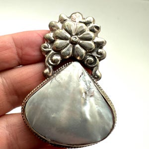 May include: A silver-toned pendant with a pearlescent, teardrop-shaped center stone. The stone is framed by a silver border and topped with a floral design. The pendant is likely a piece of jewelry.