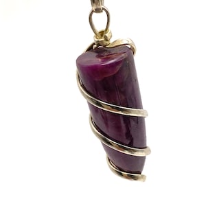 May include: A pendant featuring a polished, deep purple gemstone wrapped in silver-colored wire. The gemstone is cylindrical with a rounded top and is suspended from a silver chain. The pendant is set against a white background.