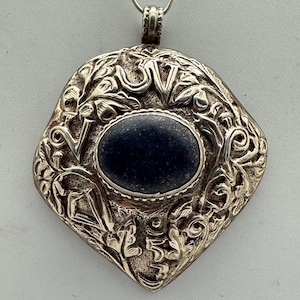 May include: Silver-toned pendant with an oval, dark blue stone at the center. The pendant has an ornate, floral design around the stone and is suspended from a thin silver chain. The pendant is approximately 2 inches in length.