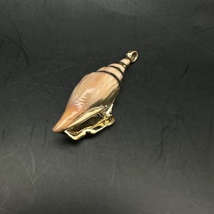 May include: A pendant shaped like a spiral seashell with a smooth, light tan exterior and a polished gold-tone metal base. The shell features a tapered top with a small, integrated gold-tone metal loop for attachment to a chain or cord.