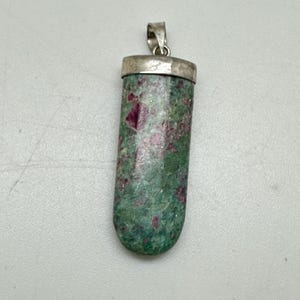 May include: A cylindrical pendant with a silver-colored bail. The pendant is a mix of green and red hues, with a polished finish. The gemstone pendant is approximately 1.5 inches long, suitable for necklaces.