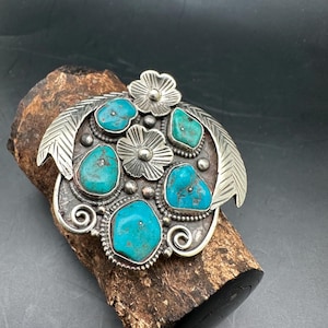 May include: A silver-toned metal belt buckle with turquoise stones and floral accents. The buckle features five turquoise stones in various shapes, surrounded by silver metal with floral and leaf designs. The buckle is resting on a piece of wood.