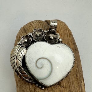 Puede incluir: Un colgante en forma de corazón con una concha de ojo de shiva blanca con un patrón de espiral natural, engastado en un marco de metal plateado. El colgante está decorado con tres pequeñas flores de metal y un motivo de hoja lateral.