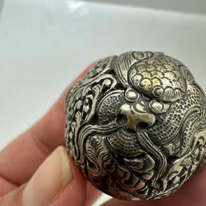 May include: A spherical metal object with a dark, tarnished silver finish featuring intricate, raised relief carvings of a dragon. The surface displays detailed scales, swirling cloud-like patterns, and a prominent dragon head with textured features.
