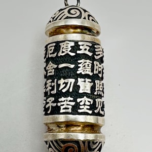 以下が含まれることがあります： 精巧なスクロール模様と漢字が施されたシルバーカラーのペンダント。円筒形の本体は黒い背景に白い文字が特徴です。上部と下部には装飾的なシルバーのデザインが施されています。チェーン用の小さなループが上部にあります。