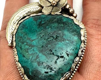 May include: A large silver ring featuring a prominent, irregular, dark teal-green stone with black matrix. The setting is decorated with a silver leaf motif on the left side and a small five-petaled flower at the top, surrounded by a beaded silver border.