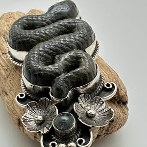 May include: A detailed pendant featuring a coiled snake design. The snake is dark gray with a textured surface, set within a silver frame. The pendant includes silver floral accents and a central gemstone, creating a unique jewelry piece.