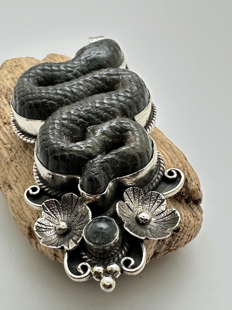 May include: A detailed pendant featuring a coiled snake design. The snake is dark gray with a textured surface, set within a silver frame. The pendant includes silver floral accents and a central gemstone, creating a unique jewelry piece.