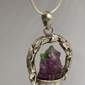 May include: A silver pendant necklace featuring a carved gemstone of a deity. The deity is purple and green, set within a textured silver frame. The necklace has a delicate silver chain.