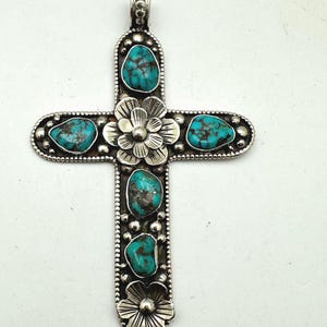 May include: Silver-toned cross pendant with turquoise stones and floral accents. The cross features five turquoise stones and decorative floral elements at the center and bottom. The pendant is approximately 3 inches long.