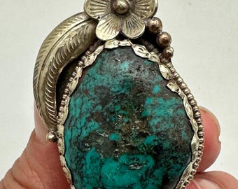 May include: A silver ring featuring a large, irregular, dark turquoise stone with black matrix. The setting is decorated with a metal flower and a curved leaf motif, surrounded by a scalloped silver border and small silver beads.