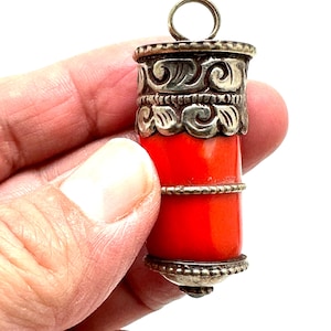 May include: A cylindrical pendant with a bright red body and ornate silver-coloured metalwork at the top and bottom. The pendant has a small loop at the top for a chain. The metalwork features a detailed, swirling design.