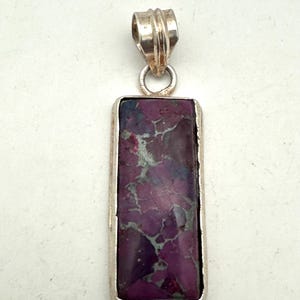 May include: A rectangular pendant featuring a polished purple stone with intricate grey veining, set in a smooth silver-toned metal frame. The pendant includes a ribbed silver-toned metal bail at the top for attachment to a chain or cord.