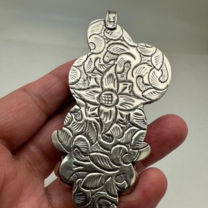 May include: A silver pendant with an intricate floral design. The pendant features a central flower surrounded by detailed leaves and petals. The metal has a slightly oxidized finish, enhancing the details.