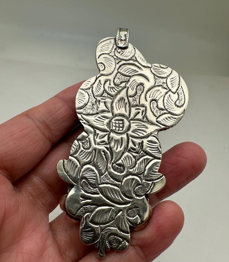 May include: A silver pendant with an intricate floral design. The pendant features a central flower surrounded by detailed leaves and petals. The metal has a slightly oxidized finish, enhancing the details.