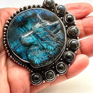 Può includere: Ciondolo argentato con una testa di lupo scolpita in pietra di labradorite blu e turchese. Il ciondolo è circondato da un bordo di perline e piccole pietre grigio scuro. Il design è circolare e dettagliato.